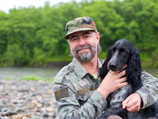 Bearded man in camouflage hat and jacket smiling and holding a dog