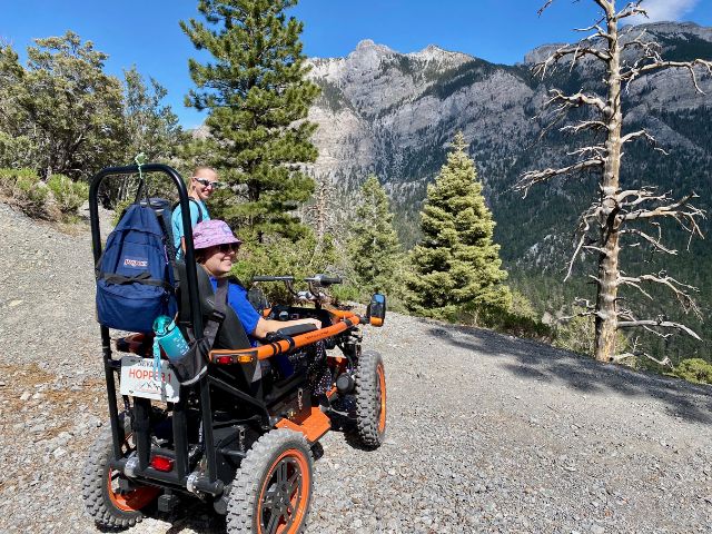 a woman in an all terrain wheelchair smiles while looking out over a desert mountain view at 9,000 ft