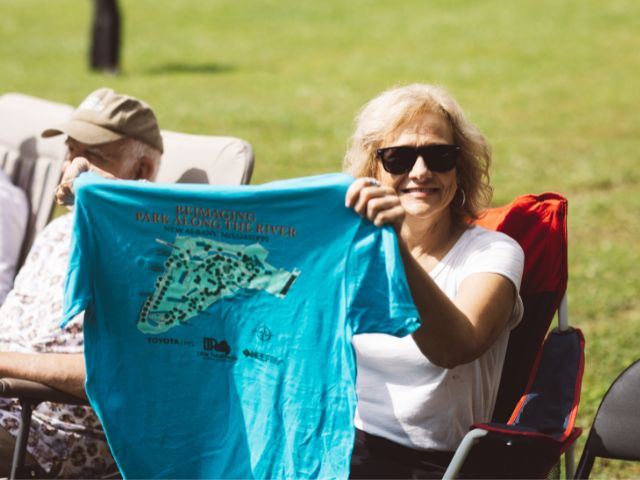 a woman holds up the back of a t-shirt with the Park Along the River revitalization plan