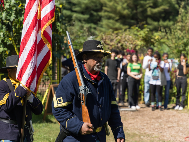 A man in a blue Union soldier uniform marches in formation as a group of people watch in the background