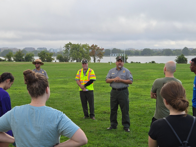 NPS Director Chuck Sams stands next to other NPS rangers as he speaks with a group of volunteers. They are standing in an open green field. Behind them in the distance is the Potomac River, with the US Capitol Dome just visible on the horizon.