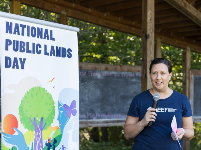 A woman wearing a blue shirt with NEEF's logo on it stands in front of a colorful banner for National Public Lands Day. She is holding a microphone and speaking to an audience off-camera.