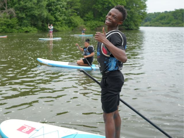 a student stands on a paddleboard smiling while on a river during a water quality and microplastics environmental education lesson.