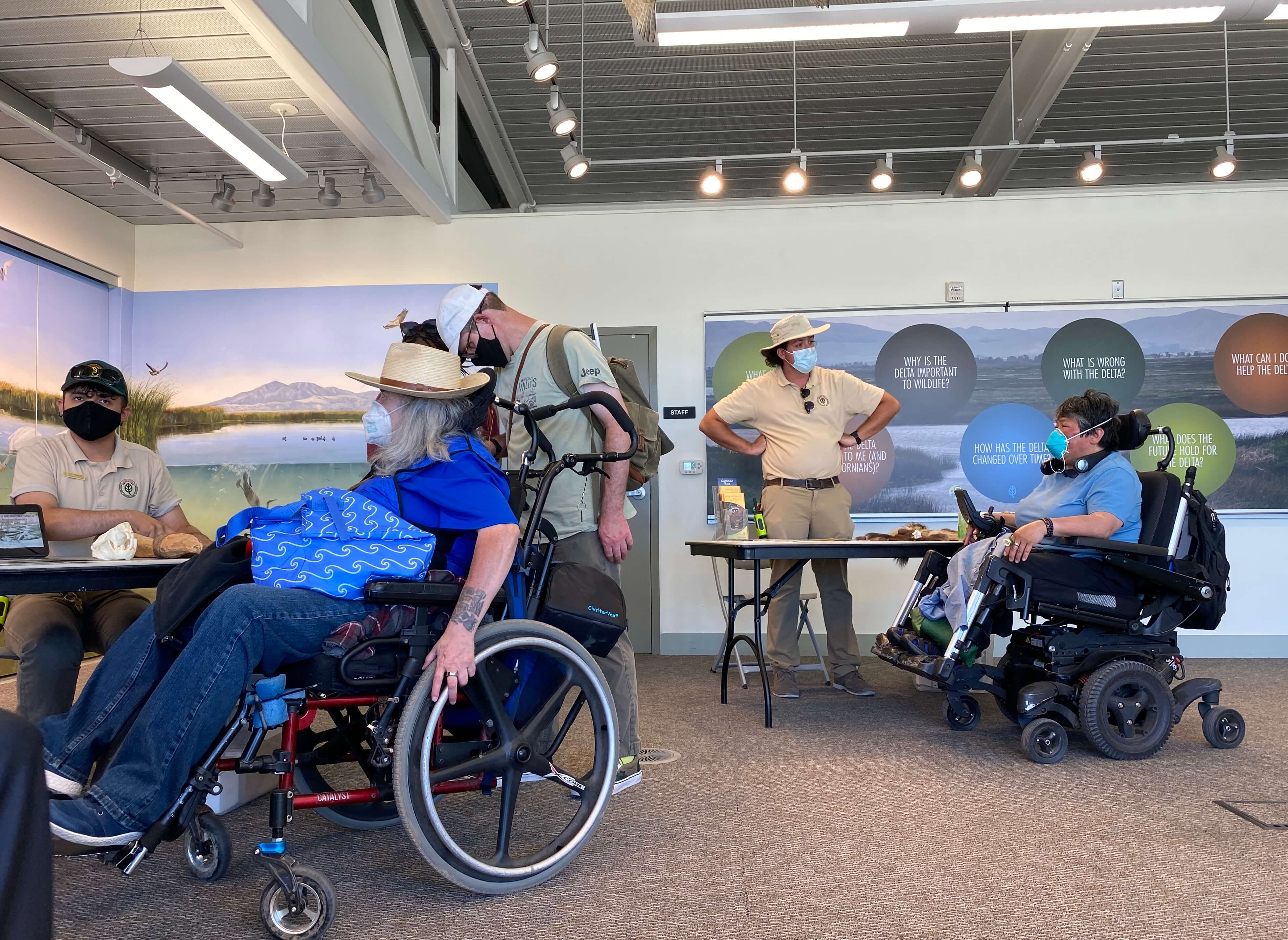 Visitor center: Park staff behind tables set with pelts and objects found in the park talk with visitors in wheelchairs