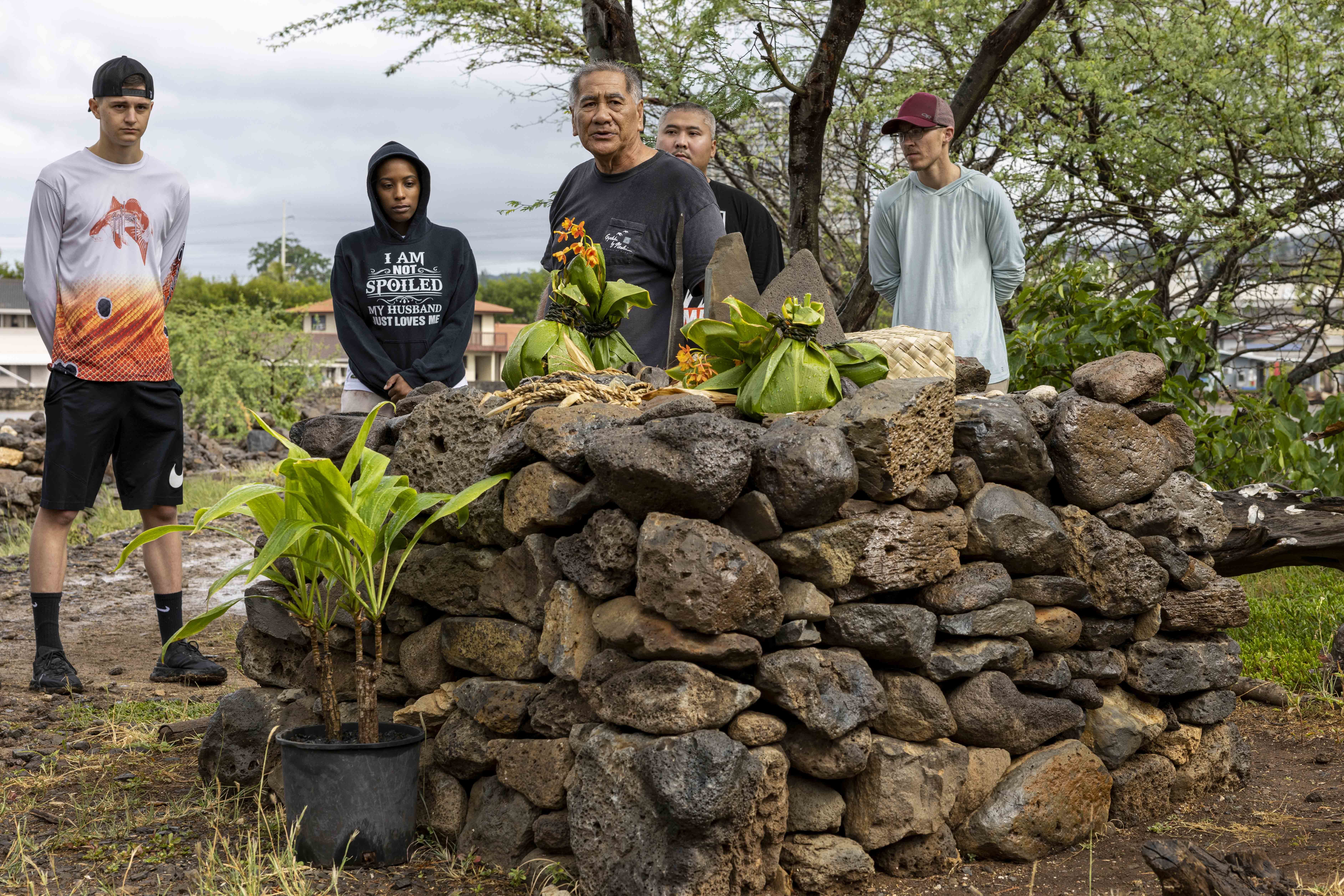 Several men stand behind a large altar consisting of stacked rough stones with traditional Native Hawaiian offerings placed on top