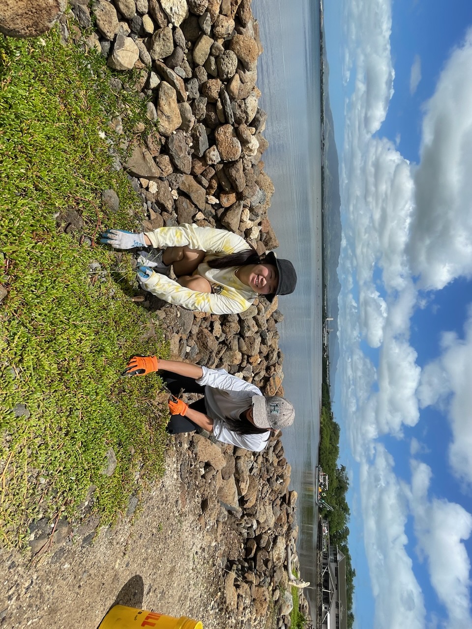 Two women are on their hands and knees plucking out invasive plants from the ground. A large body of water is behind them.