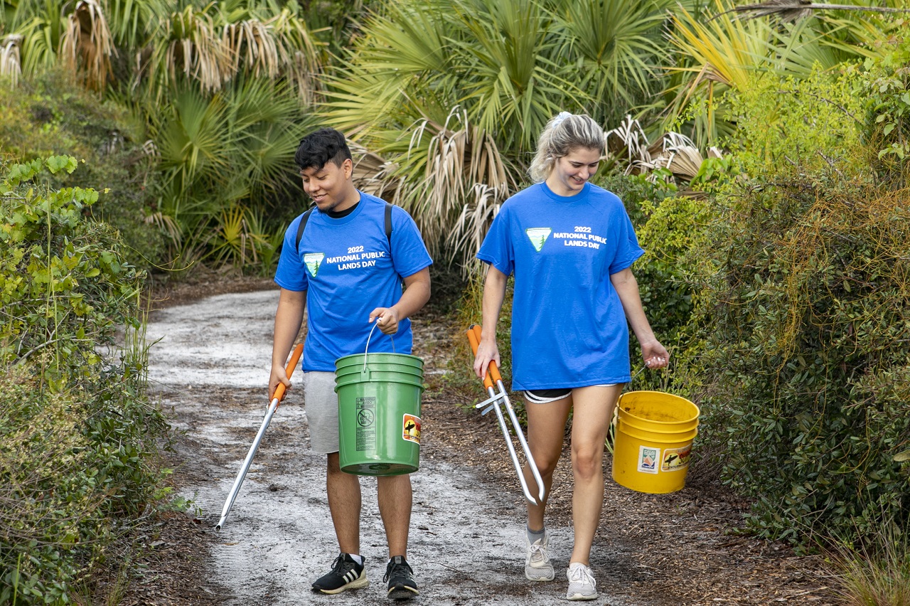 Two young people using pickers to remove trash in a green, tropical park