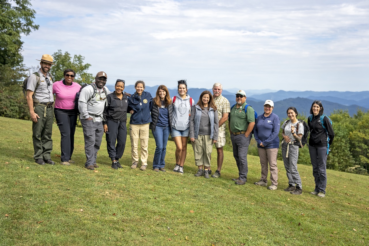 Line of 8 people dressed in outdoor gear standing on a hill with mountains in the background