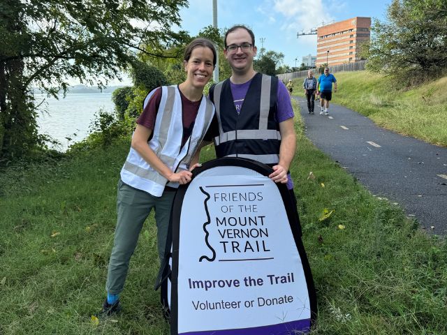 A couple stands together behind a sign advertising the Friends of the Mount Vernon Trail. They are wearing bride- and groom-themed high visibility vests.