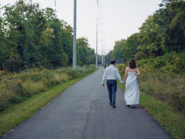 A bride and groom, facing away from the camera, walk hand-in-hand down a paved nature path surrounded by greenery