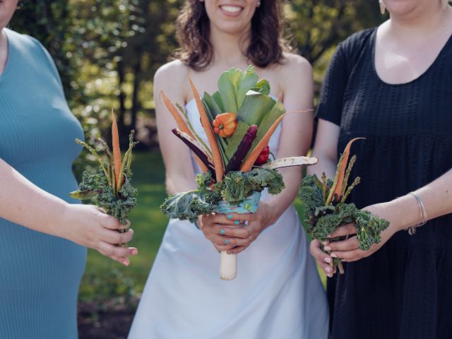 Three women stand while holding out custom wedding bouquets made from peppers, carrots, leeks, and other root vegetables.