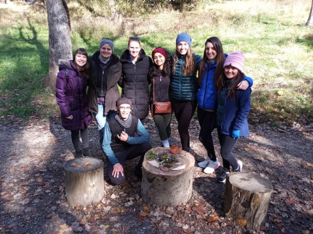 A group of young women wearing warm athletic gear pose with a man squatting by a couple of tree stumps in a forested area.