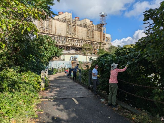 A group of volunteers work to remove invasive kudzu vines that have completely covered the fencing along a paved multi-use path. A decommissioned old power plant looms in the background.