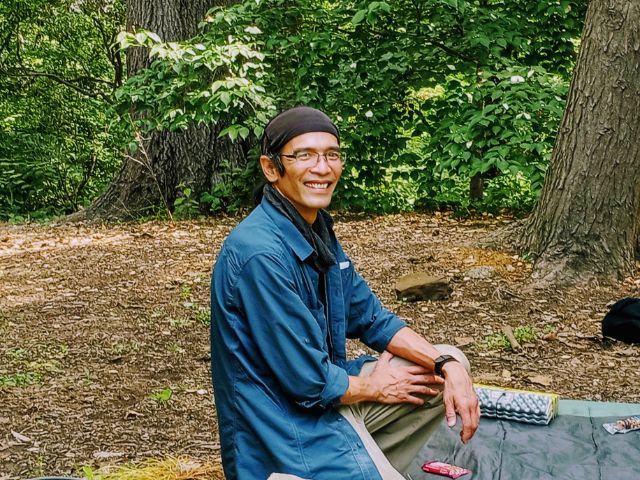 A man wearing a stocking cap, glasses, blue shirt, and tan pants poses for a photo at a forested campsite.