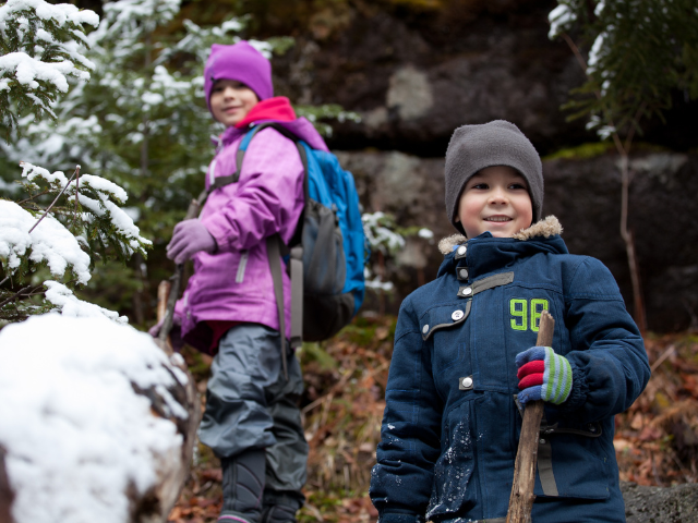 Two young children wearing winter coats and hats hike in a forest with snow all around