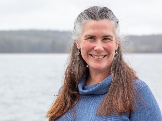 A woman with gray hair wearing a light blue sweater stands in front of an overcast, rocky shoreline