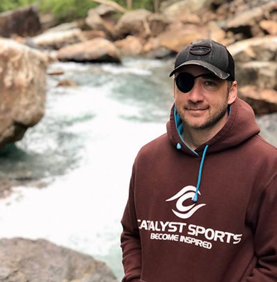 A man wearing a hooded sweatshirt, camo-print baseball cap, and an eyepatch over one eye stands in front of a waterfall.