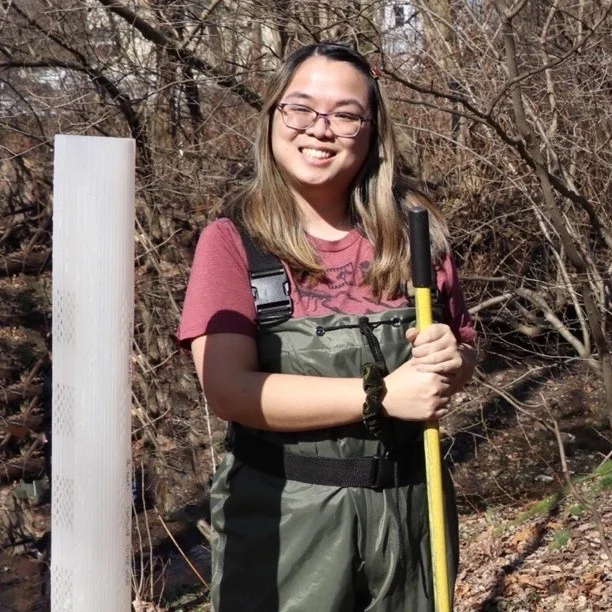 A woman with long brown hair and glasses wearing waders and holding a shovel stands near a creek 