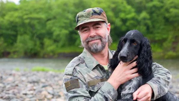 a man in army fatigues holds a dog while outside by a river bank