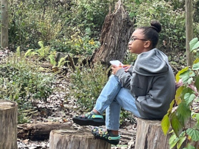 A young woman sits on a stump in a wooded area. She is holding a pad of paper and a pencil and is staring pensively into the distance.