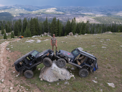 A man stands on top of two off-road vehicles parked on a large boulder. A vast mountain range extends behind him