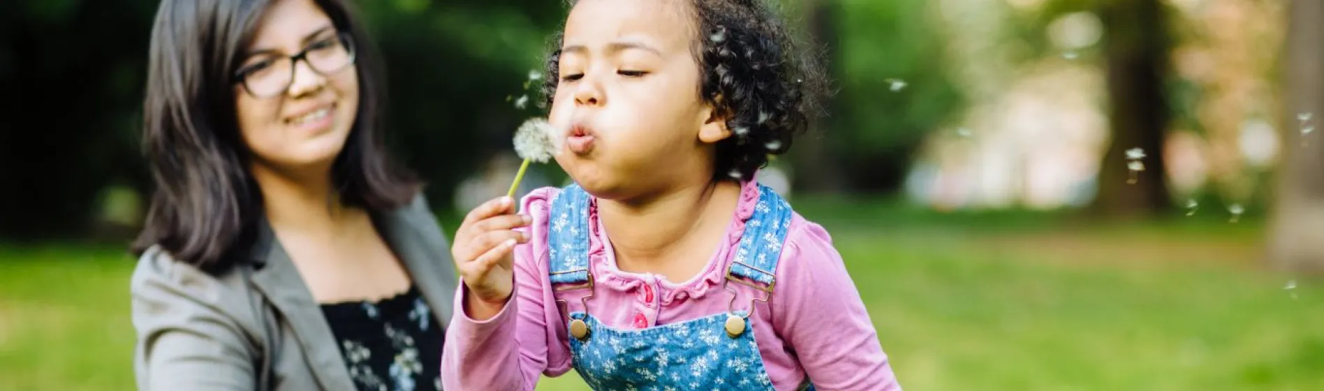 a young girl blows on a dandelion with her mother in the background