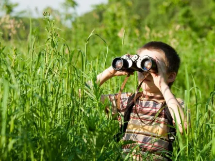 a young boy stands in a tall grass field looking through binoculars