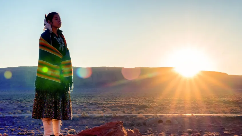 Native American young woman wearing traditional dress stands in front of a desert mountain range with sun setting.