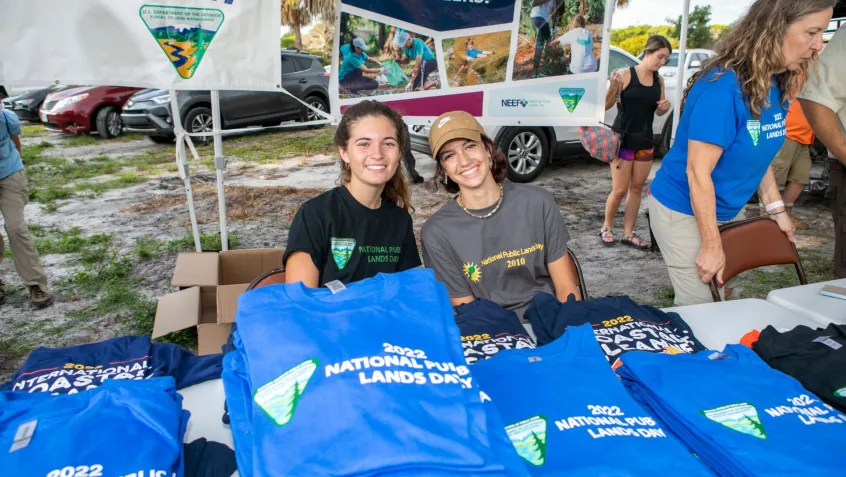 two young women sit at a NPLD event welcome table filled with National Public Lands Day tshrits