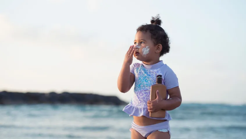 a little girl applies sunscreen to her face while standing on the beach