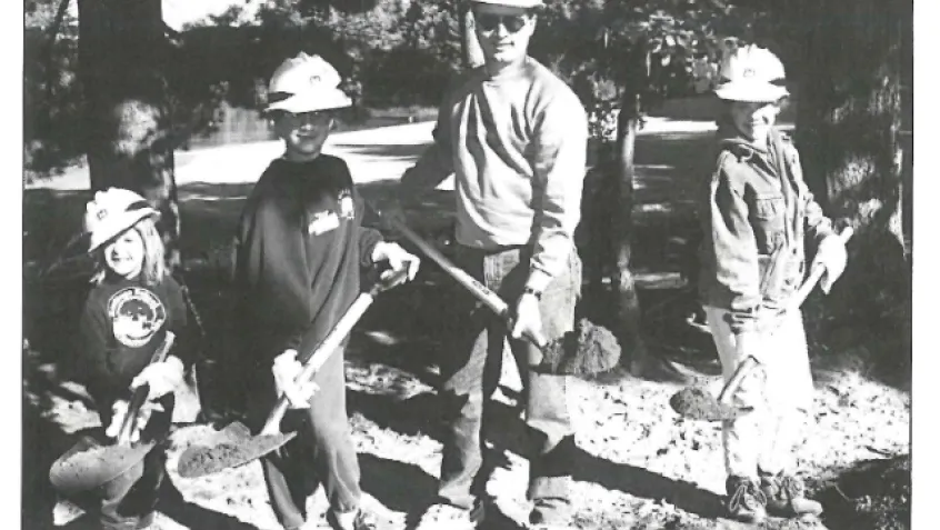 One adult and three children in hard hats with shovels outdoors smiling