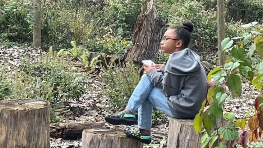 A young woman sits on a stump in a wooded area. She is holding a pad of paper and a pencil and is staring pensively into the distance.