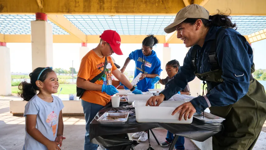 A woman shows a young girl how to perform a water quality monitoring activity at an NPLD volunteer event. Several young people are working on the project at a table in the background.