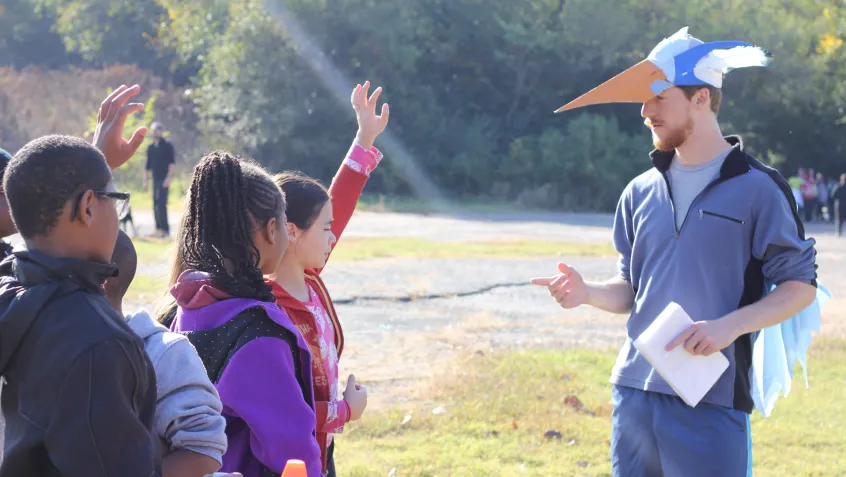 A man wearing a handmade bird hat instructs students while standing in an open field. The students are raising their hands