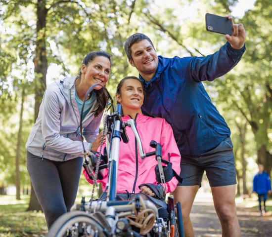 a woman sitting on a wheelchair bike take a selfie with a man and woman standing next to her.