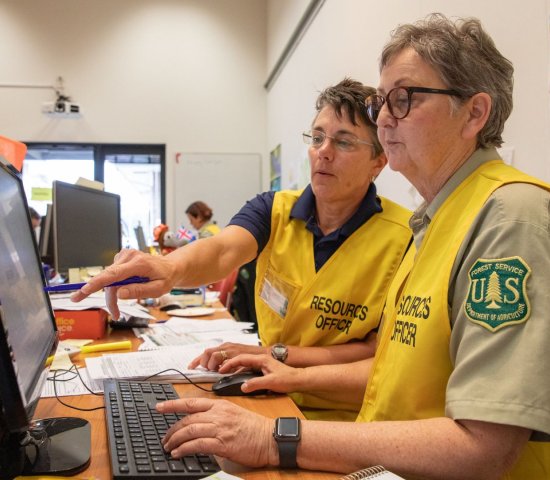 Two women wearing US Forest Service uniforms work together at a desk