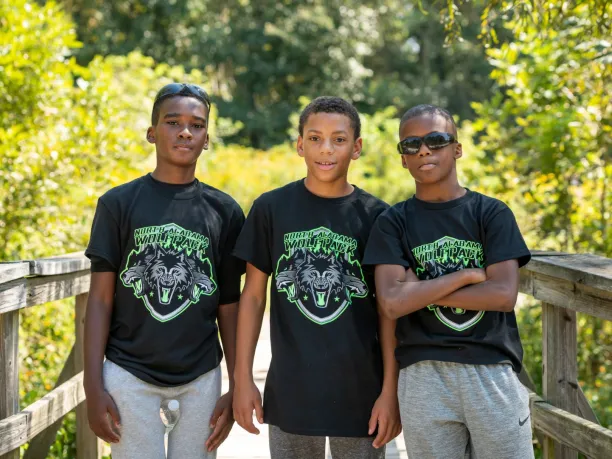 Three young black men stand together on a boardwalk in nature.