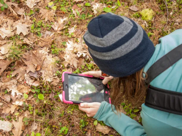 person with long hair wearing hat taking photo with phone of leaves on the ground