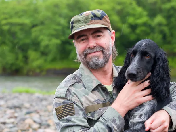 a man in army fatigues holds a dog while outside by a river bank