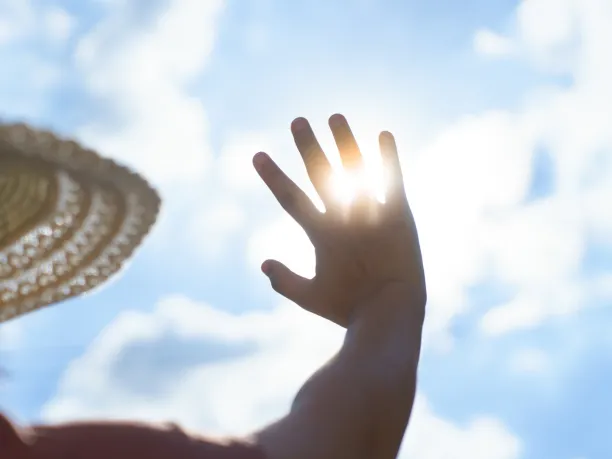 A person wearing a large sun hat holds their hand up to the sky to block the sun's harsh rays.