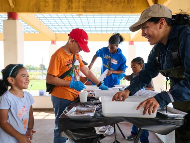 A woman shows a young girl how to perform a water quality monitoring activity at an NPLD volunteer event. Several young people are working on the project at a table in the background.