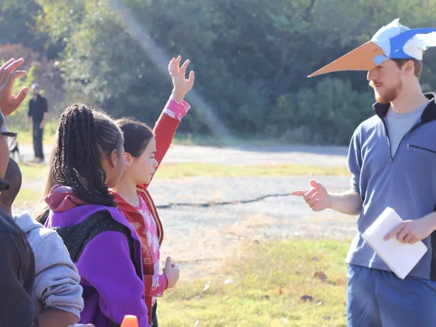 A man wearing a handmade bird hat instructs students while standing in an open field. The students are raising their hands