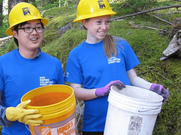 Young woman and man holding buckets and wearing hard hats volunteering outdoors