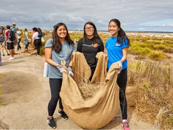 Three young women pose for a photo holding a large bag of invasive plants they've removed from a local habitat.