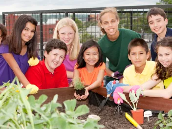 A diverse group of students pose for a photo in front of a raised garden bed in a school garden. A young girl in the center is holding a small plant that is about to be planted in the bed.